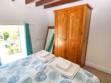 A bedroom with a wooden wardrobe a standing mirror a bed with patterned bedding and folded towels near a window with blue curtains at Newton West Farm Newton near Newgale