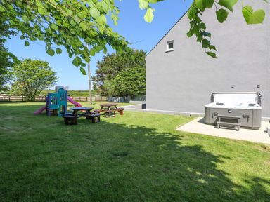 A backyard with a play structure, picnic tables, and a hot tub next to a gray building at Newton West Farm in Newton near Newgale