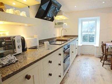 A kitchen with countertop appliances and cabinets next to a window with a dining table and chairs at Beach Cottage in Little Haven Pembrokeshire