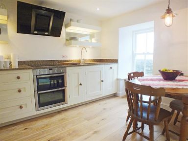 A kitchen with white cabinets a double oven a sink wooden dining table and chairs a bowl of fruit and a window at Beach Cottage in Little Haven Pembrokeshire