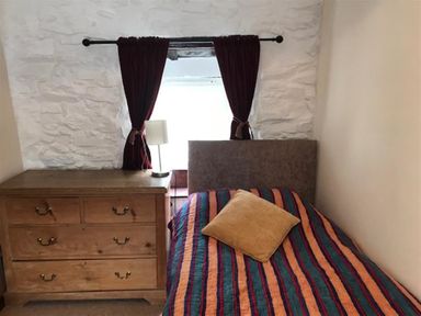 A bedroom with a bed covered by a striped blanket and a chest of drawers with a table lamp next to a window with curtains at Beach Cottage in Little Haven Pembrokeshire