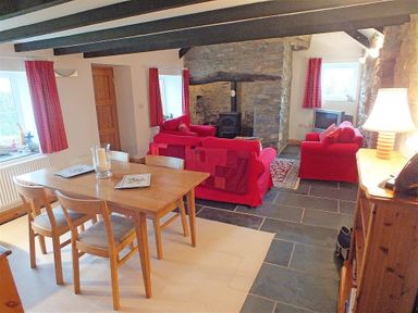 A dining and living area with a wooden table and chairs red sofas a stone fireplace and exposed wooden beams at Gwndwn Gwyn in Trefasser Near Strumble Head