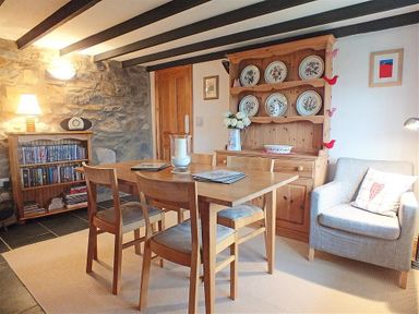 A dining area with a wooden table and chairs a wooden cabinet with decorative plates a wooden bookshelf with books a gray armchair with a cushion and a stone wall at Gwndwn Gwyn in Trefasser Near Strumble Head