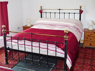 A bedroom with a black metal bed frame a striped bedspread and red blanket two wooden bedside tables with lamps and red curtains at Gwndwn Gwyn in Trefasser Near Strumble Head
