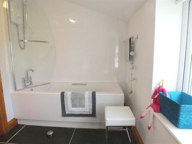 A bathroom with a bathtub that has a shower and a towel hanging on it a white stool a blue basket and a doll on the window sill at Gwndwn Gwyn in Trefasser Near Strumble Head