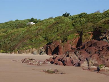 A sandy beach with reddish rocks and green foliage on a hillside with a house at Beach Cove in Freshwater East Pembrokeshire
