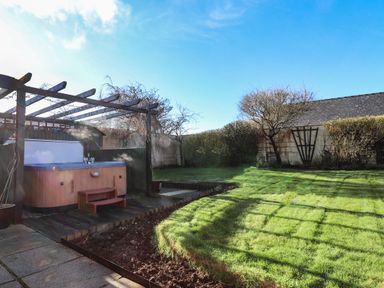 A backyard with a hot tub under a pergola on a wooden deck and a grassy lawn surrounded by trees and bushes at Y Stabl in Fishguard Pembrokeshire