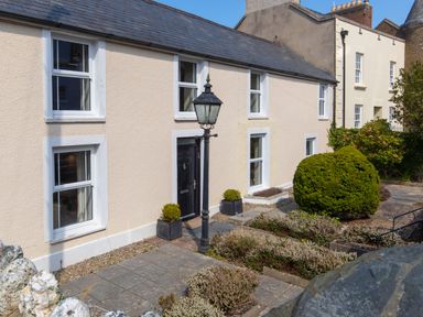 Front exterior of a beige house with six white-framed windows a black door a black lamp post two potted plants and garden shrubs at The Rock House in Fishguard Pembrokeshire