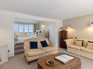 A living room with two beige plaid sofas and a wicker coffee table connected to a sunken seating area with white armchairs and a view of the sea at The Rock House in Fishguard Pembrokeshire