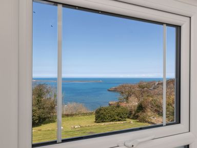 A window view of the sea with a lawn and bushes in the foreground and trees on the right at The Rock House in Fishguard Pembrokeshire