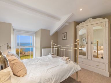 A bedroom with a metal bed frame white bedding an antique wardrobe with mirrors and a window with a sea view at The Rock House in Fishguard Pembrokeshire