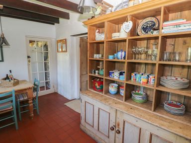 A kitchen with a wooden cupboard filled with dishes and cups and a wooden table with chairs near a glass paneled door at Ty-Newydd Cottage in Nevern near Newport Pembrokeshire