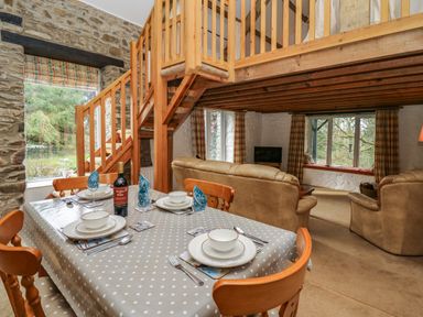 A dining table with place settings and a bottle of wine next to a wooden staircase and a living area with sofas and large windows at Trawsnant Cottage in New Quay Ceredigion