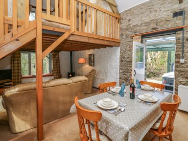 A dining area with a set table and wooden chairs next to a living room with sofas and a wood stove under a wooden mezzanine at Trawsnant Cottage in New Quay Ceredigion