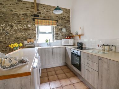 A kitchen with wooden cabinets and tiled floor a stone wall with window and a plaid curtain at Trawsnant Cottage in New Quay Ceredigion