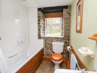 A bathroom with a bathtub a toilet with a wooden seat and a window with stone walls at Trawsnant Cottage in New Quay Ceredigion
