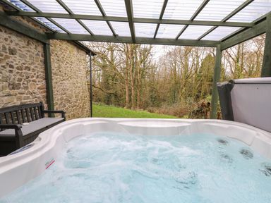 A bubbling hot tub with a bench beside a stone wall under a transparent roof overlooking trees at Trawsnant Cottage in New Quay Ceredigion