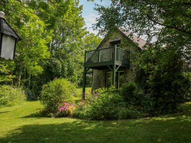 A stone cottage with a wooden balcony surrounded by green trees and garden at Trawsnant Cottage in New Quay Ceredigion