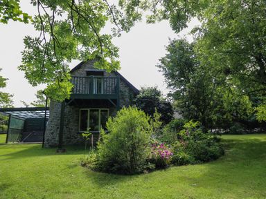 A stone house with a balcony surrounded by trees and a garden with flowers at Trawsnant Cottage in New Quay Ceredigion