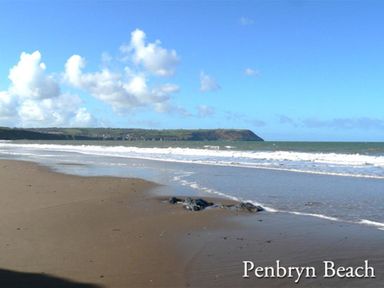 A sandy beach with waves and rocky coastline under a blue sky at Penbryn Beach