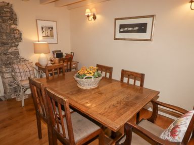 A dining room with a wooden table and six chairs a basket with flowers on the table a side table with a lamp and books and framed pictures on the walls at 1 Cheriton Row Carew Cheriton Nr Tenby Pembrokeshire