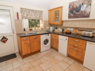 A kitchen with wooden cabinets a washing machine a dishwasher a toaster and a window with a floral blind at 1 Cheriton Row Carew Cheriton Nr Tenby Pembrokeshire