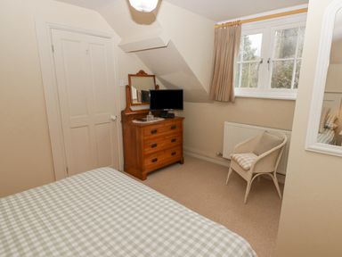 A bedroom with a bed covered in checkered bedding a wooden dresser with a mirror and a television a white chair with cushions under a window at 1 Cheriton Row Carew Cheriton Nr Tenby Pembrokeshire