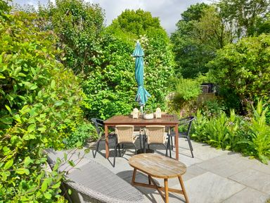 A garden patio with a wooden table, chairs, an umbrella, and a small wooden coffee table surrounded by green plants and trees at 1 Cheriton Row Carew Cheriton, Nr Tenby, Pembrokeshire