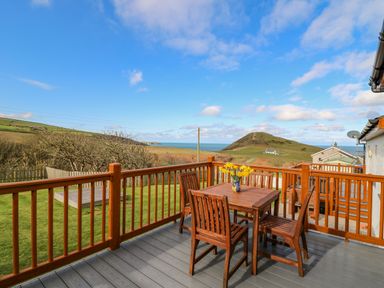 An outdoor deck with a wooden table and four chairs with a vase of yellow flowers overlooking hills and the sea at Llaethdy in Cardigan