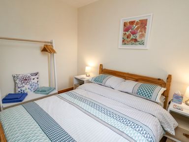 A bedroom with a wooden bed, patterned bedding, bedside tables with lamps, a floral picture on the wall, and a clothing rack with hangers at Llaethdy in Cardigan