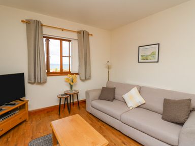 A living room with a beige sofa with cushions a wooden coffee table a television on a wooden stand a window with curtains and small nesting tables with flowers at Beudy in Mwnt Cardigan Ceredigion