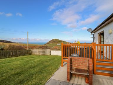 A backyard with green grass a wooden fence a brick barbecue and a wooden deck attached to a white house with a view of hills and the sea at Beudy in Mwnt Cardigan Ceredigion