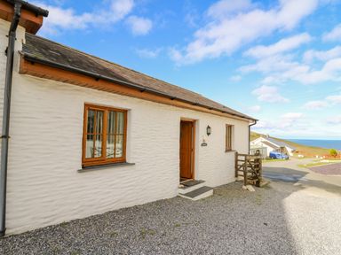 Exterior of a white stone cottage with wooden window frames and an open door on a gravel driveway with a view of other houses and the sea at Beudy in Mwnt Cardigan Ceredigion