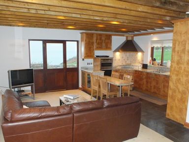 A kitchen and living area with a brown leather sofa wooden dining chairs a television and wooden cabinets at Cable Cottage in Fishguard Pembrokeshire