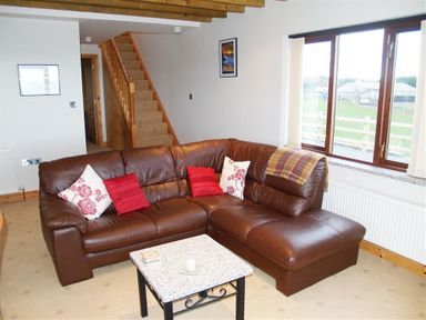 A living room with a brown leather sectional sofa with cushions a small table stairs and window with a view at Cable Cottage in Fishguard Pembrokeshire