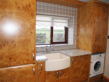 A kitchen area with wooden cabinets a white farmhouse sink a washing machine and a window with a roller blind at Cable Cottage Fishguard Pembrokeshire