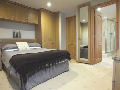 A bedroom with a double bed grey bedding and a black throw built in wooden wardrobes a door leading to a bathroom with a glass shower enclosure