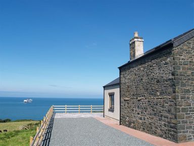A stone and plaster house with a chimney near a gravel driveway overlooking the ocean with a ship in the distance at Cable Cottage Fishguard Pembrokeshire