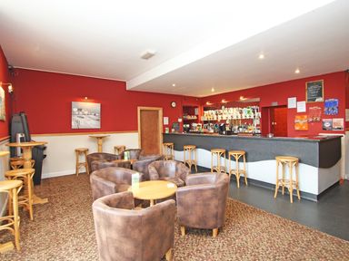 An interior of a bar with a long counter and bar stools surrounded by round tables and brown upholstered chairs at 9 Manorcombe Bungalows St Ann's Chapel near Callington