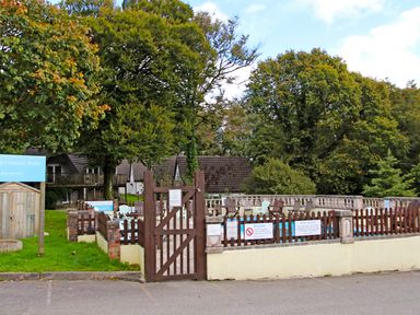 An outdoor swimming pool area with a wooden gate and fence surrounded by trees and cottages at 9 Manorcombe Bungalows in St Ann's Chapel near Callington