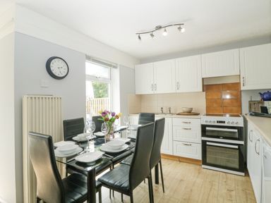 A kitchen with a glass dining table set with plates bowls and glasses four black chairs a window white cabinets an oven and a clock on the wall at 9 Manorcombe Bungalows in St Ann's Chapel near Callington