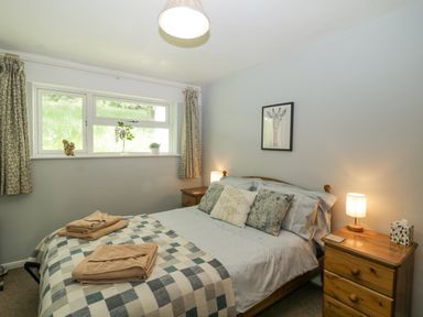 A bedroom with a double bed with three pillows a checkered blanket and towels on top two wooden bedside tables with lamps a window with curtains and plants on the sill and a framed giraffe drawing on the wall at 9 Manorcombe Bungalows in St Ann's Chapel near Callington