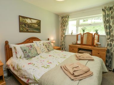A bedroom with a wooden bed and floral bedding a dressing table with mirrors and patterned curtains at 9 Manorcombe Bungalows in St Ann's Chapel near Callington