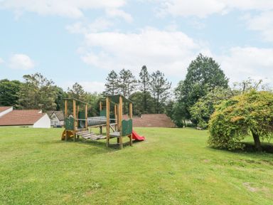 A playground with slide and climbing structure on a lawn with houses and trees at 9 Manorcombe Bungalows in St Ann's Chapel near Callington