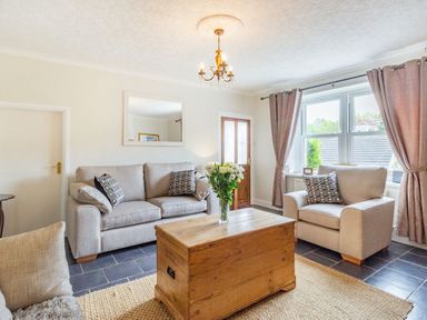 A living room with beige sofas a wooden chest as a coffee table flowers on the table a window with curtains and a chandelier at Peth Head Cottage in Wooler