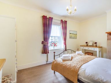 A bedroom with a bed covered with a yellow quilt and white towels by a window with red curtains and a dressing table with a mirror at Peth Head Cottage in Wooler