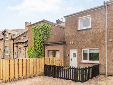 The exterior of a brown stone house with a small black fence and a wooden fence on the side at Peth Head Cottage in Wooler