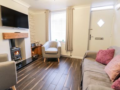 A living room with a wood stove fireplace armchairs a sofa with cushions a wall-mounted television and a front door at Scarborough Cottage in Driffield
