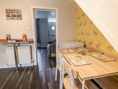 A dining area with a wooden table and chairs next to a wall with rabbit patterned wallpaper and a small counter with books near a doorway showing a glimpse of a sitting room at Scarborough Cottage in Driffield