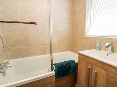 A bathroom with a bathtub, a sink with a wooden cabinet, a towel hanging on the tub, and a window with blinds at Scarborough Cottage in Driffield
